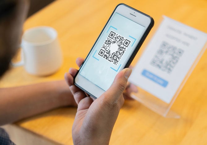 Mercado Pago: imagem das mãos de uma pessoa segurando um celular e apontando a tela para um QR Code impresso em uma mesa que pode representar um QR Code falso colocado no lugar do original por golpistas para induzir o consumidor a ações que podem levar ao roubo de dados sensíveis.