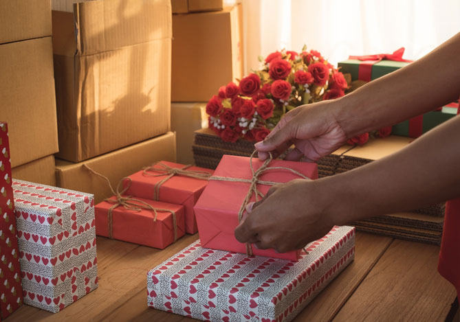 Mercado Pago: mesa com caixas e presentes embrulhados com papel estampado de coração, além de flores vermelhas e mãos de uma pessoa embrulhando um presente após validar ideias para o Dia dos Namorados