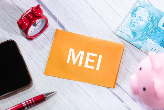 Mercado Pago: mesa vista de cima, com itens espalhados por ela, sendo um papel escrito “MEI”, uma caneta, um celular, um despertador, um cofrinho e uma nota de cem reais. Tudo isso representando o pagamento do imposto MEI.