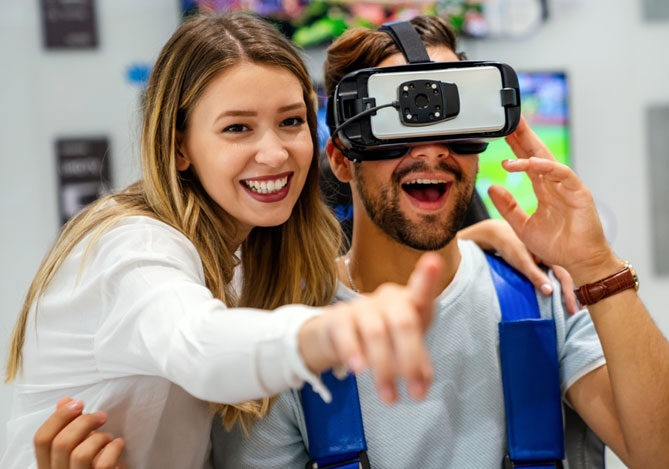 Mercado Pago: imagem de um casal sorrindo. Na imagem é possível ver o rapaz experimentando um óculos de realidade virtual em uma loja de videogames enquanto a mulher sorri e aponta com a mão direita para o que pode ser uma tela em frente aos dois.