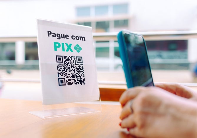 Mãos de uma mulher segurando o celular e apontando ele para o cartão impresso com o QR Code para a pessoa pagar com Pix, sugerindo que agora é possível pagar com Pix ao aproximar o celular na maquininha Mercado Pago