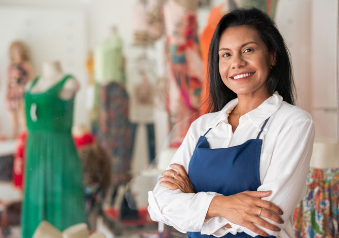 Mercado Pago e mês das mulheres: mulher de camisa branca e avental azul em primeiro plano sorrindo e com braço cruzado, ao fundo, manequins com roupas coloridas, sendo uma loja de roupas.