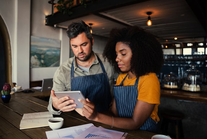 Mercado Pago: homem e mulher de avental no balcão de um bar olhando planilhas em um tablet para definir as metas SMART do seu negócio. 