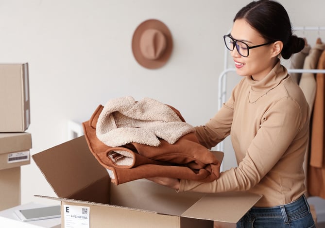 Mercado Pago: empreendedora encaixotando um casaco marrom e felpudo da sua loja que foi vendido na campanha de moda inverno e será enviado para a cliente. 