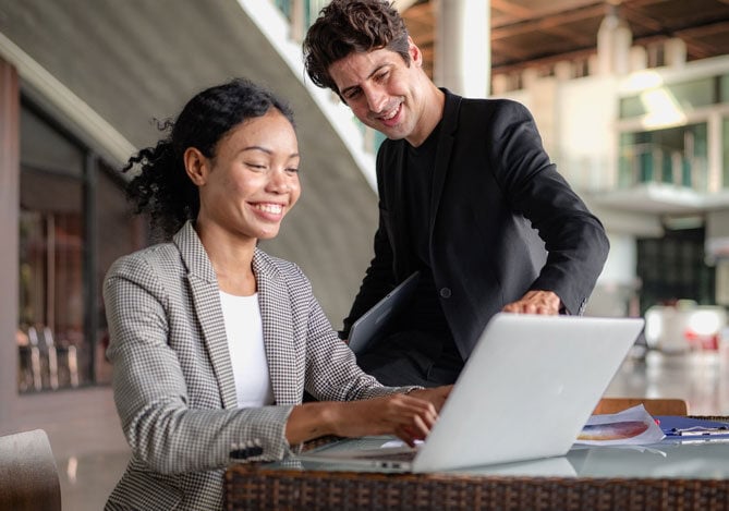 Mercado Pago: imagem de uma mulher sentada à mesa de trabalho, enquanto digita em um notebook para pesquisar sobre modelos de negócio para empreender. Ao seu lado, há um homem sorrindo a ajudadando a escolher entre as diversas opções, a que melhor se encaixa na realidade dela.