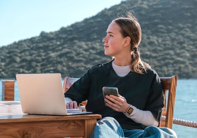 Mercado Pago: imagem de uma pessoa sentada à beira mar - aparentemente em um deck de madeira - digitando em um notebook com uma das mãos, enquanto segura um celular com a outra mão, ilustrando que essa pessoa está fazendo a gestão de informações sobre o modelo de negócio que ela escolheu para fazer uma renda extra durante suas férias. 