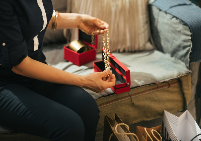 Mercado Pago: vendedora de joias com roupa preta sentada em sofá com colar dourado na mão realizando suas suas vendas com NFC no celular. Apoiado no sofá, há duas caixas vermelhas de joias e sacolas no chão.