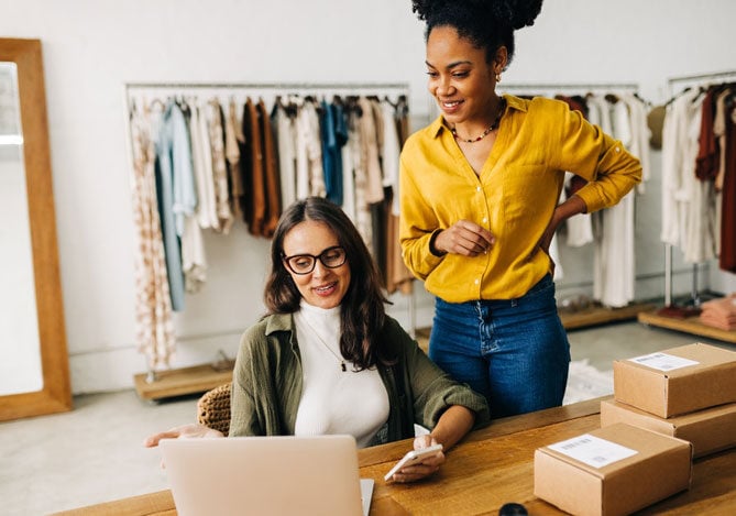 Empreendedoras analisando estratégias para explorar um nicho de mercado em uma loja de roupas, com caixas prontas para envio e computador em uso.