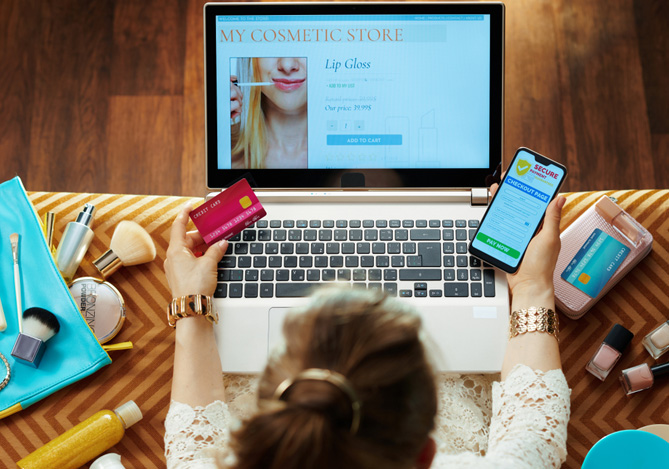 Mercado Pago: mulher fotografada de cima, em frente a um notebook com tela aberta na loja de produtos, enquanto ela segura um cartão em uma mão e um celular em outra. Ao redor, produtos de beleza para ela fazer a revenda de cosméticos.