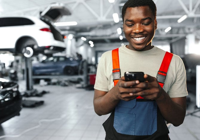 Mercado Pago: trabalhados de oficinas mecânicas no seu ambiente de trabalho, mexendo no celular com um sorriso no rosto para acessar a entrada dos pagamentos recorrentes. Atrás dele, um carro erguido para manutenção e mais alguns veículos no pátio.