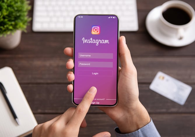 Pessoa acessando a tela de login para entrar em seu perfil do Instagram em um smartphone, com mesa de trabalho ao fundo contendo café, caderno e cartão.