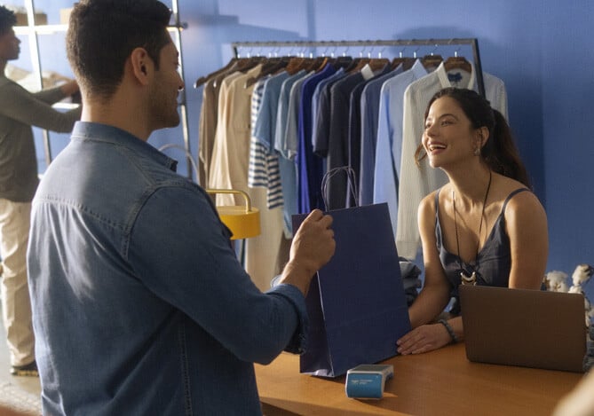 Empreendedora entregando a sacola com a compra feita por um homem vestindo camisa jeans e no balcão a maquininha Point Mercado Pago.