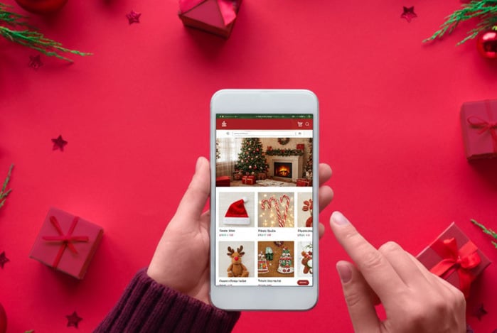 Mercado Pago: mãos segurando um celular visto de cima em frente a um fundo rosa com presentes. A tela está em um e-commerce de produtos de Natal para comprar online. 