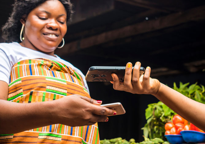 Mercado Pago: imagem de uma empreendedora ao receber por aproximação pelo celular em seu comércio. Na imagem, é possível ver que a empreendedora está segurando seu celular para cobrar pela venda, enquanto a cliente paga usando seu celular. ao fundo da imagem, é possível ver verduras e legumes, indicando que a venda é em uma feira.