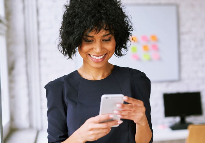 Mercado Pago: mulher com roupa casual em um escritório home office olhando sorridente para a tela do seu celular para decidir se utiliza o lucro da sua empresa para reinvestir no negócio ou criar uma reserva.