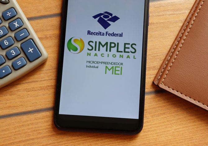 Mercado Pago: imagem de um celular sobre uma mesa, entre uma calculadora e uma carteira de couro. Na tela do celular há o logo do app MEI, ilustrando a responsabilidade fiscal do empreendedor em acessar o aplicativo e cumprir com suas obrigações para manter o MEI regular. 