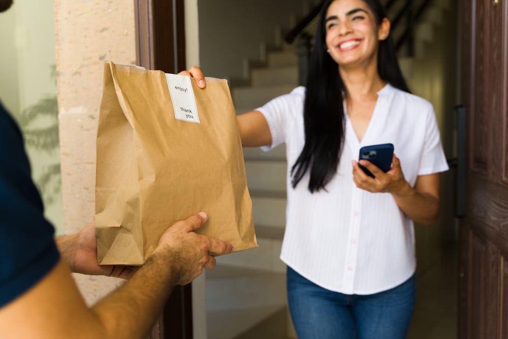 Mercado Pago: mulher de cabelo preto longo sorrindo e vestindo blusa branca e calça jeans segurando o celular com uma mão e na outra pegando a comida que pediu no restaurante delivery das mãos do entregador.