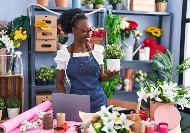 Mercado Pago: Mulher em sua floricultura com avental azul e blusa branca, segurando um vaso com uma planta. Ao redor, há serviços e produtos que ela oferece, como arranjos e itens como papéis e fitas para pacotes.