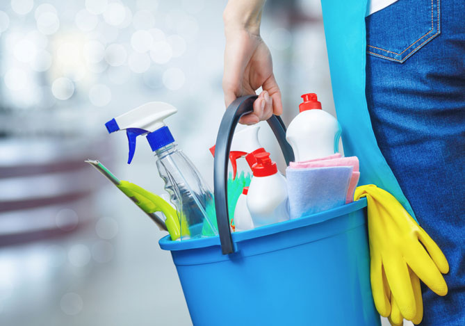 Mercado Pago: Pessoa com calça jeans e avental azul de costas que presta serviços de limpeza segurando balde azul com luvas amarelas e diversos produtos de limpeza dentro do balde, como sprays, rodo e panos.