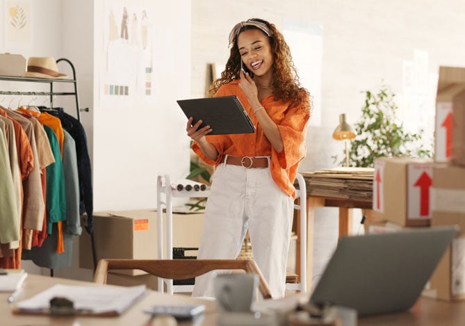 Mercado Pago: imagem de empreendedora vestindo calça branca e camisa laranja em seu escritório. Na imagem, é possível perceber que ela está falando ao telefone, enquanto segura, em uma das mãos, um tablet no qual acompanha os dados de venda e estoque de seu negócio por meio de um sistema integrado.