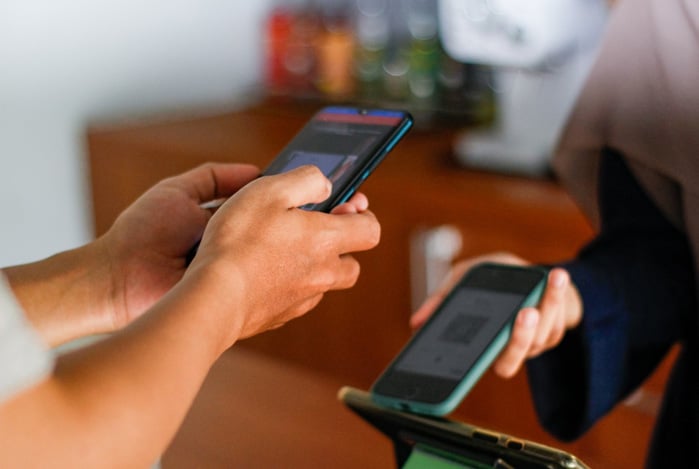 Mão de um vendedor segurando o celular com a tecnologia Tap to Pay no iPhone a partir do Mercado Pago para cobrar o cliente que também segura seu celular para efetuar o pagamento da sua compra. 