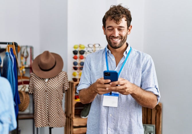 Mercado Pago: homem de crachá e celular na mão, dentro da sua própria loja de roupas, sorrindo enquanto solicita a antecipação de recebíveis.