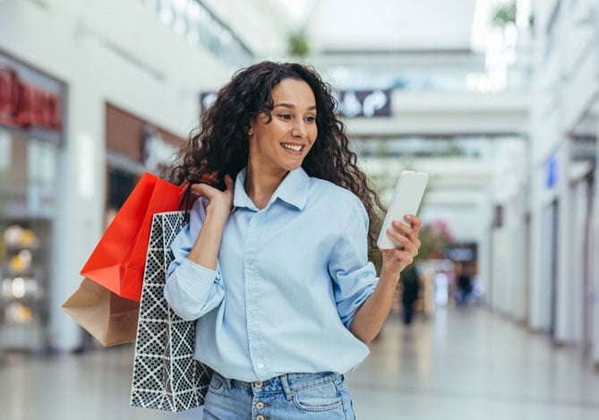 Mercado Pago: imagem de uma consumidora vestindo camisa azul e calça jeans e caminhando pelo corredor de um shopping com algumas sacolas de compras, enquanto olha para a tela do seu celular sorrindo. A imagem ilustra uma consumidora que está finalizando uma compra online pelo dispositivo móvel e aproveitou uma oportunidade de sugestão de compra de venda cruzada para adquirir mais produtos no checkout e economizar no frete.