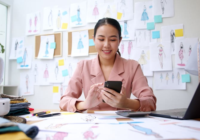 Mercado Pago: empreendedora de costura em seu atelier, com máquina em cima da mesa e vários desenhos colados na parede, enquanto ela está sentada à mesa com um notebook e seu celular na mão para realizar as vendas digitais das peças produzidas. 