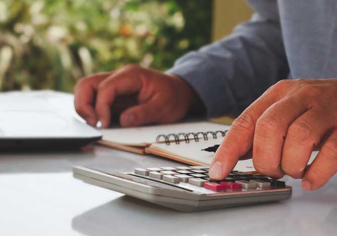 Mercado Pago: Imagem ilustrativa de mão de homem digitando em calculadora na mesa de escritório, com caderno e notebook, realizando imposto PJ.