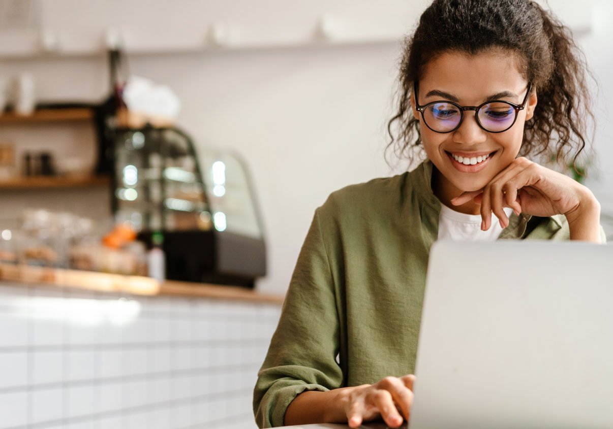 Mulher de óculos sorrindo e sentada enquanto mexe no seu laptop