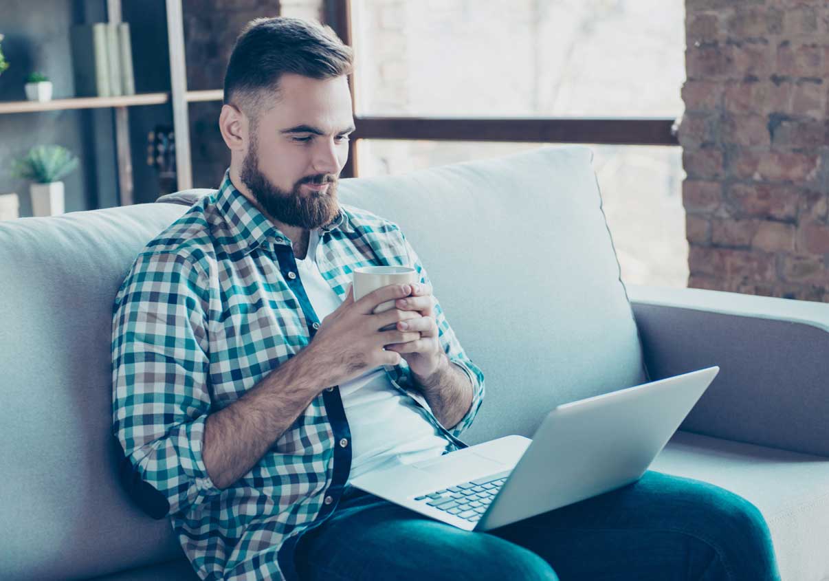 Homem sentado no sofá com o notebook no colo e segurando uma xícara 