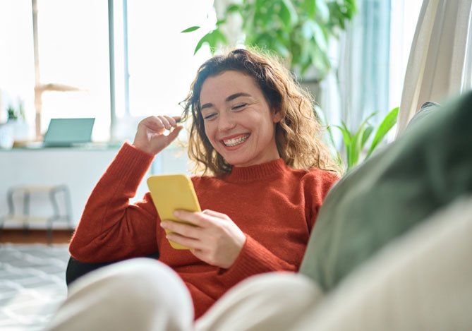Mercado Pago: Empreendedora feliz sentada em seu sofá usando o celular para medir a satisfação do cliente de seu negócio.