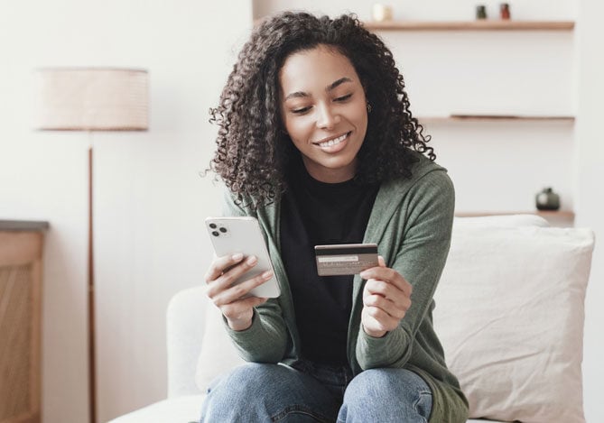 Imagem de uma mulher segurando um celular em uma das mãos e um cartão na outra para realizar um pagamento parcelado via Link de Pagamento Mercado Pago que recebeu por meio de aplicativo de mensagens. A mulher está sentada em uma poltrona e veste calça jeans azul, camiseta preta e casaco verde. 